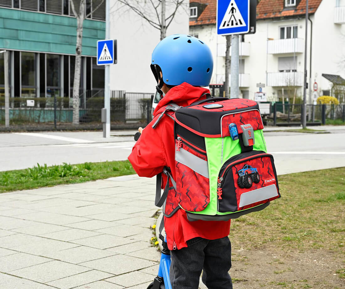 Junge fährt mit einem Cityroller an der Straße und trägt einen roten Grundschulranzen auf dem Rücken mit Monstertruckmotiv